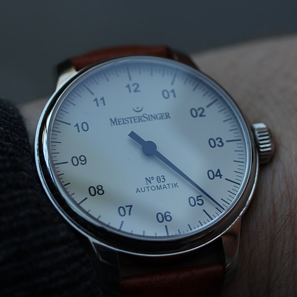 MeisterSinger microbrand watch with single hand, worn on man's wrist, dark grey background.