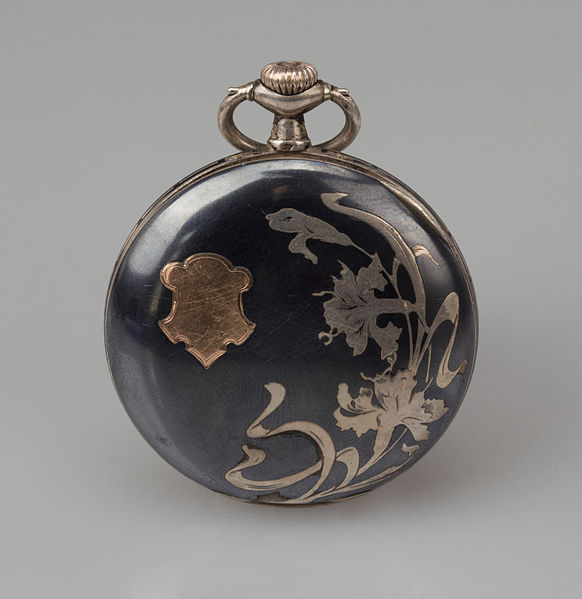 Art Nouveau watch decoration on pocket watch, dark with paler metal decorations, on grey background.