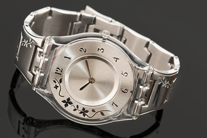 Silver coloured watch featuring its metal surfaces, with decoration and metal strap, on dark reflective surface.