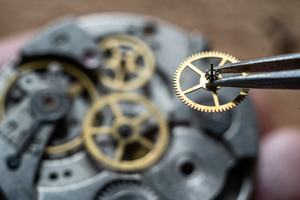 Watch assembly, showing a gear being held in metal tweezers over a complex watch mechanism, shown out-of-focus.