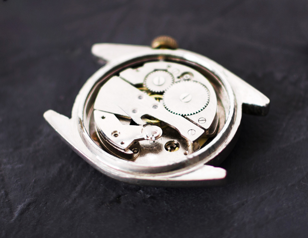The inside of a watch showing gears and mechanism in a metal case, on a dark grey textured stone surface.