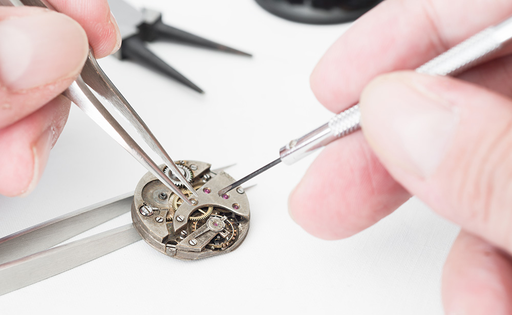 Watch assembly, showing man holding tools and a watch movement with gears and purple jewels.