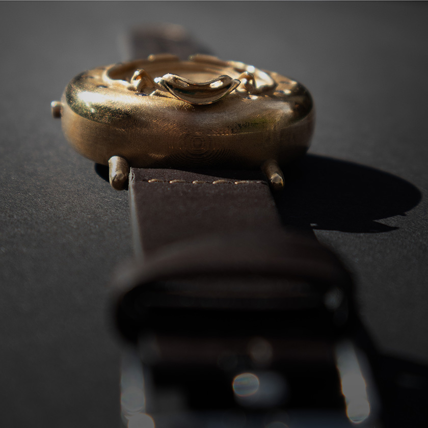 UnconstrainedTime Fractal Emergence wristwatch in bronze, shallow angle end view on dark paper, with brown leather strap.